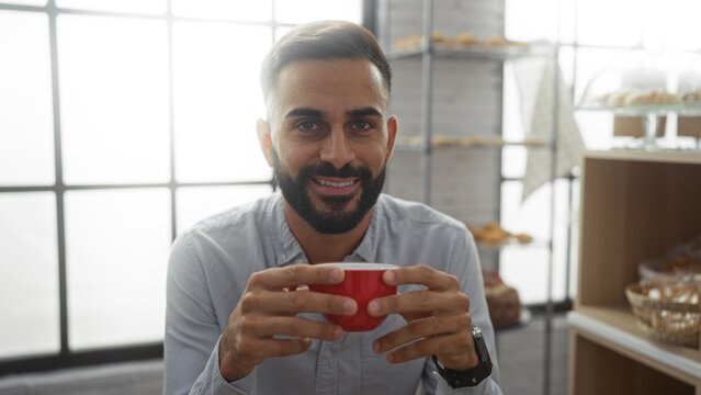 Handsome young hispanic man enjoying a coffee in a cozy bakery with a bright interior, seated indoors holding a red cup and smiling at the camera
