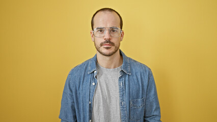 Portrait of a confident hispanic man with a beard, glasses and denim shirt against a yellow...