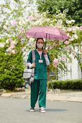 Nurse standing outdoors with purple umbrella, wearing green scrubs and carrying a bag. Vibrant flowering trees and green foliage in background creating contrast