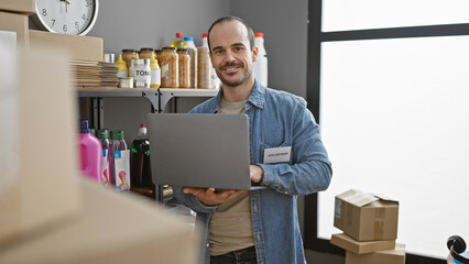 Smiling bald man with a beard using a laptop in a warehouse full of donation boxes and supplies,...