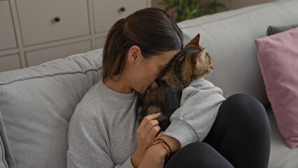 A young hispanic woman cuddling her cat on a cozy sofa in a modern living room, demonstrating a loving bond between pet and owner indoors.