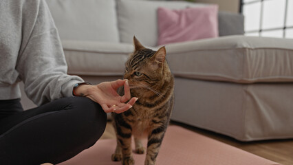 A hispanic woman in the living room of her home gently touches a tabby cat while sitting on the floor indoors. © Krakenimages.com