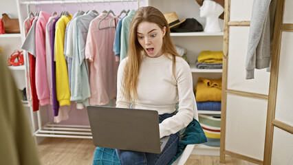 Astonished young woman with blonde hair using laptop in a fashion wardrobe room.