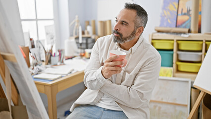 Mature bearded man contemplates in studio, holding cup, surrounded by art supplies, embodying creativity and introspection.