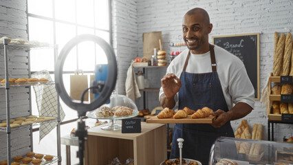 Young man recording a video in a bakery shop while holding a plate of croissants, surrounded by various baked goods and shelves, under warm indoor lighting.