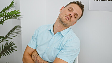 A tired young man with a beard and tattoo sleepily awaits in a clean, minimalist room, signaling a...