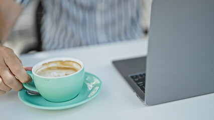 Close-up of a caucasian male's hand holding a coffee cup on a terrace with a laptop in the background