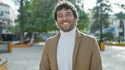 Fototapeta premium A smiling young hispanic man with a beard stands casually outdoors in a city park, exuding a relaxed and handsome demeanor.