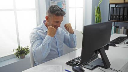 Naklejka premium Handsome man celebrating success at his office desk indoors, emphasizing a positive workplace environment