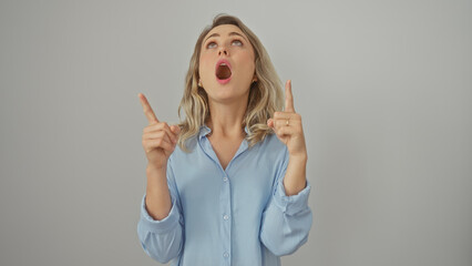 A surprised young woman with blonde hair pointing upwards, isolated against a white background, expressing astonishment.