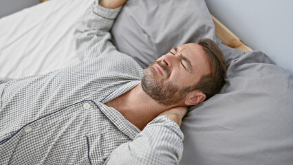 Middle-aged hispanic man with a beard in pain, lying in bed at home, clutching his neck.