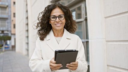 A smiling middle-aged hispanic woman in a white coat using a tablet on a city street.