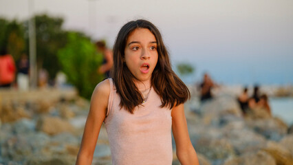 A dark-skinned teenage girl sitting on the rocks by the seaside on a sunny summer day, showing a frightened expression