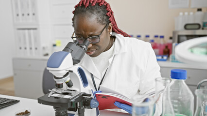 African female scientist with braids at microscope in laboratory holding book, indoor portrait