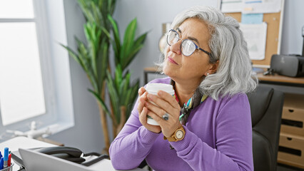 A thoughtful senior woman enjoys a coffee break at her modern office, displaying professionalism and tranquility.