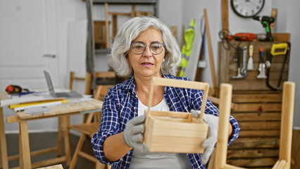 A mature woman examining a wooden crate in a well-equipped carpentry workshop indoor.