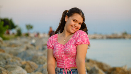 A chubby teenage girl sitting on the rocks by the seaside on a sunny summer day, smiling and looking at the camera