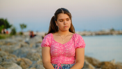 A somewhat chubby teenage girl sitting on the rocks by the seaside on a sunny summer day, showing sad facial expressions