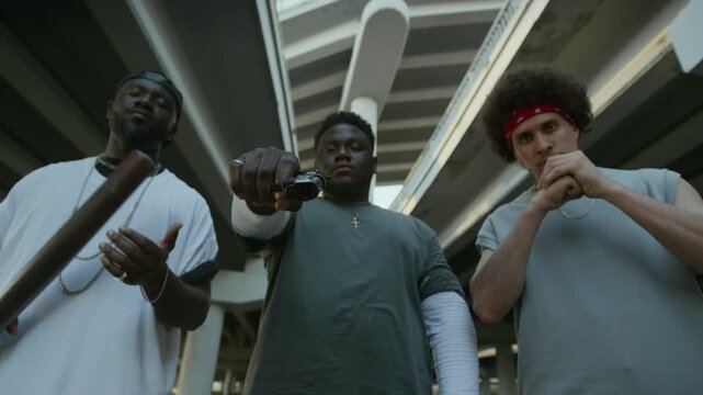 Low angle group portrait shot of criminal gang posing together on city street. Black man pointing gun at camera, his friends holding baseball bat and knuckling up