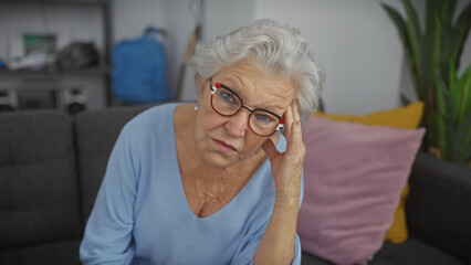 Worried senior woman with glasses seated on a couch in the living room of a modern apartment