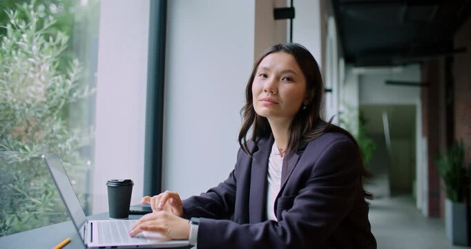 Camera slowly moving towards cute Asian woman sitting in front of her laptop in corridor. Typing on keyboard. Entering message, Looking at camera and smiling. Wearing business outfit.