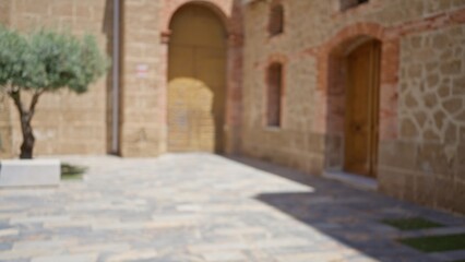 Blurred image of a quiet outdoor courtyard with stone buildings and an olive tree, featuring a rustic archway and paved flooring in a sunlit mediterranean town.