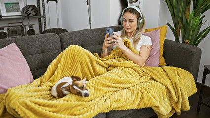 A young woman with headphones using a smartphone cuddles with her dog on a couch covered with a...