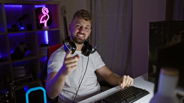 Smiling caucasian man with beard and blue eyes wearing headphones in a dark gaming room at night