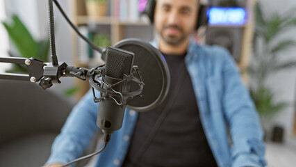 Hispanic man with beard in studio for podcast recording, sitting by microphone and mixing console.