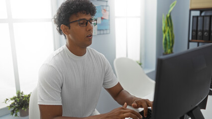 Hispanic man working on a computer in a modern office with indoor plants and a large window, focused on typing and wearing glasses and a white shirt.