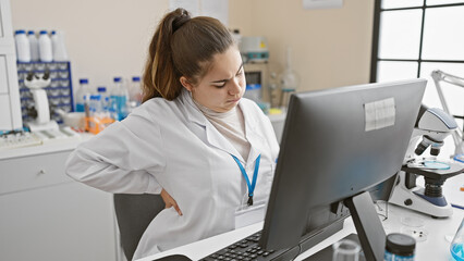 A young hispanic woman in a lab coat suffers back pain while working on a computer in a laboratory setting