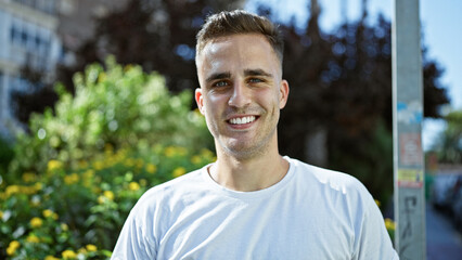 A smiling young hispanic man in a white shirt stands outdoors in a sunny park with greenery.