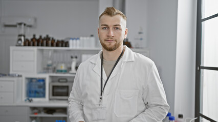 A young bearded man wearing a lab coat stands confidently in a modern laboratory setting with...