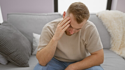 A distressed young caucasian man with a beard sitting on a gray couch indoors appears contemplative or worried.