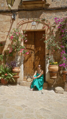 Young woman sitting in front of a rustic doorway surrounded by colorful flowers on a sunny day in valldemossa, mallorca, spain