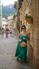 Naklejka premium Woman wearing sunglasses and holding a straw bag, walking along a rustic street in valldemossa, mallorca, spain, surrounded by historic stone buildings with a backdrop of mountains.