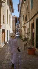 Woman walking in a quaint stone-paved street of fornalutx, mallorca, spain, dressed in a white outfit and hat on a sunny day, showcasing the charming architecture and serene atmosphere of the island.
