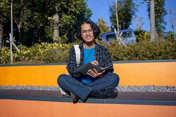 A male student is sitting in the park and writing on a book.