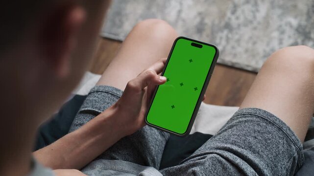 Caucasian teenager uses mobile phone with chroma key mockup, brows the Internet or scrolls social network while sitting on sofa. Teen boy spending time at home. Screen content. Green screen. Close Up.