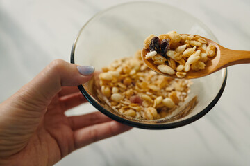 Hand holding a wooden spoon filled with mixed nuts above a transparent bowl, showcasing a variety of nuts and raisins for a healthy snack option