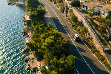  A sporty black car with a SUP on the roof races along a coastal road, the sun shining brightly overhead. 