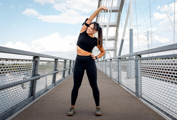 Young fitness woman in black sport outfit preparing for a run stretching her muscles at bridge.