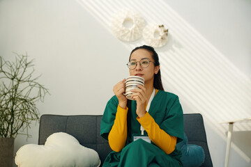Nurse sitting on couch, holding coffee mug, and gazing thoughtfully. Background includes wall decor...