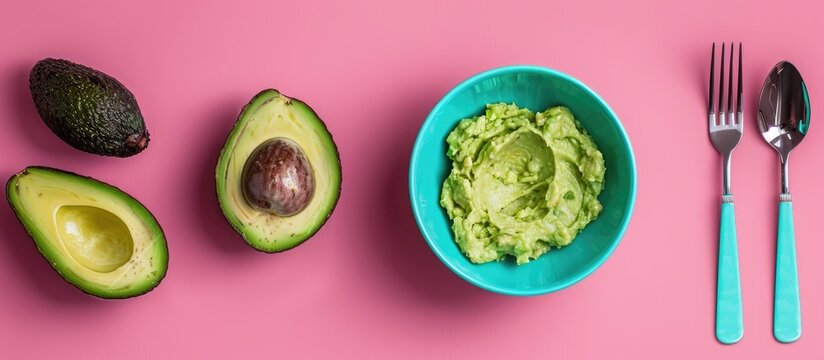 An overhead shot showing a halved Chilean Hass avocado with the pit mashed avocado in a turquoise bowl with a fork and spoon set on a pink backdrop with room for text. Copy space image