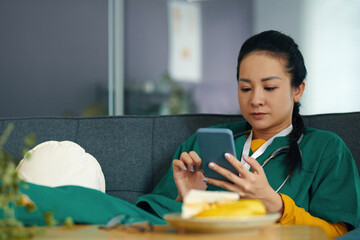 Asian nurse wearing green scrubs engrossed in reading messages on smartphone while sitting in...