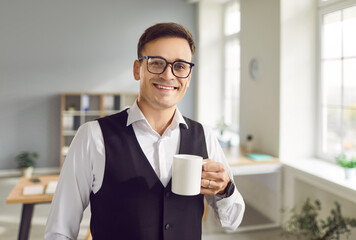 Front view portrait of a young smiling businessman, with a cup of tea or coffee. Positive, successful manager holding white mug, drinking hot beverage in modern office, looking at camera.