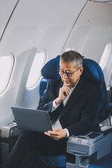 Fototapeta premium Attractive Asian male passenger of airplane sitting in comfortable seat while working laptop and tablet with mock up area using wireless connection. Travel in style, work with grace.