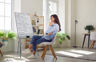 Side view portrait of young business woman sitting in office or at home in front of white board with daily plans and tasks. Girl looking at work schedule planning appointment for week.