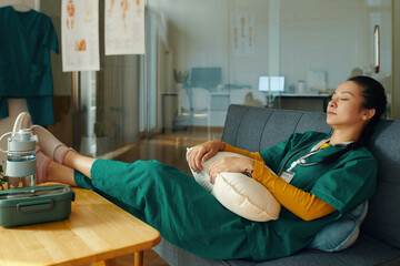 Female nurse in green scrubs taking well-deserved rest on couch in hospital lounge Nursing tools and medical posters are visible in background, indicating healthcare setting