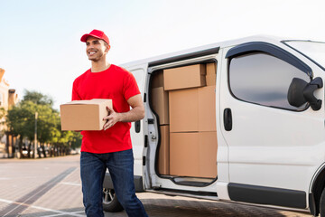 Cheerful young delivery man in red uniform carrying package cardboard box from his white van,...